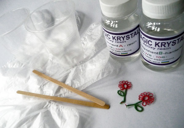 Resin casting set with tatting lace flowers: resin and hardener bottles, small plastic cups, wooden sticks, and transparent gloves, part of Tatting in resin tutorial by FairyLacefromElena.