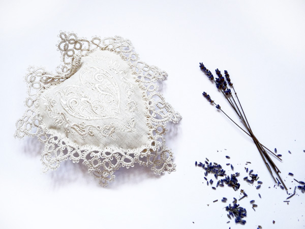 The photo shows a beige heart-shaped sachet with embroidery and hand-tatting lace. A bouquet of lavender lies nearby. The heart and lavender are two delicate symbols of love and France, illustrating the article "Cherbourg Umbrellas" by FairyLavefromElena.